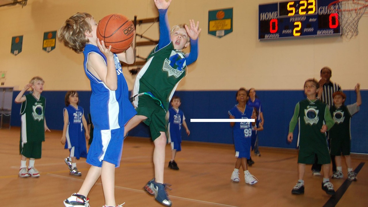 Youth basketball game in an indoor gym; a player attempts a shot while another jumps to block, with other children and a scoreboard behind them.